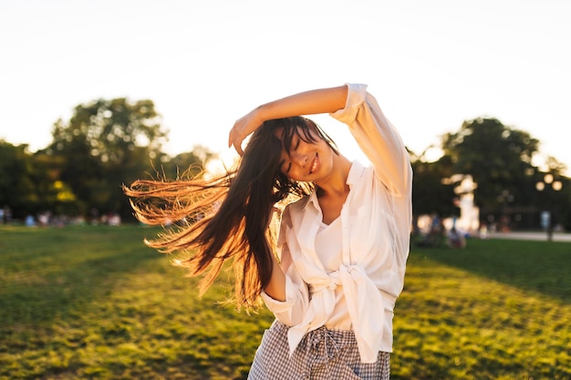 Linda garota asiática sorridente na camisa branca dançando alegremente enquanto passa o tempo no parque da cidade