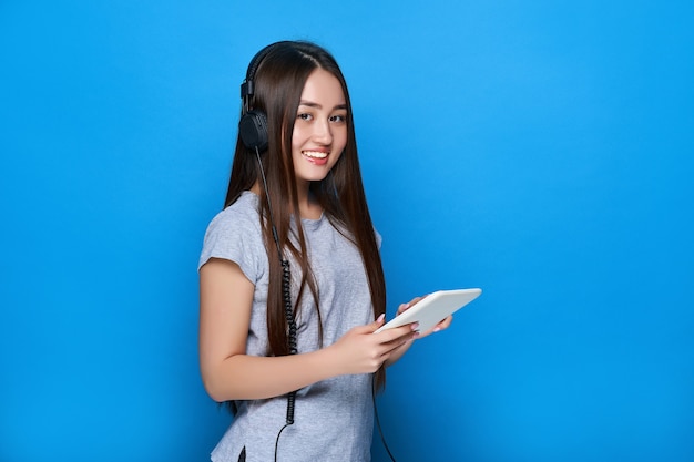 Linda garota asiática sorridente, ouvindo música em fones de ouvido e segurando o tablet na parede azul