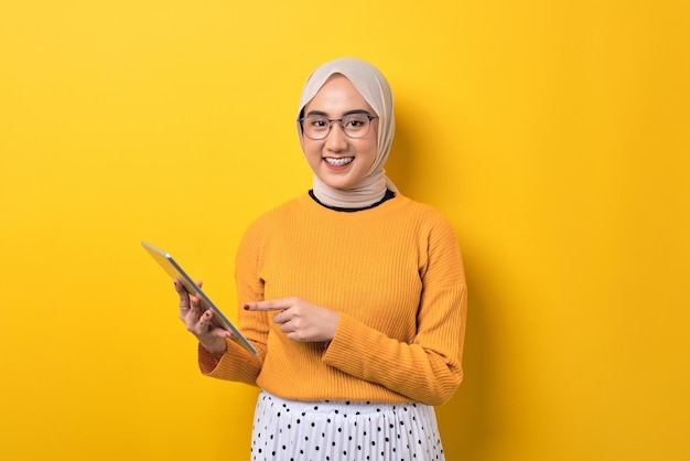 Linda garota asiática sorridente usando hijab apontando para tablet digital verificando novos aplicativos isolados em fundo amarelo