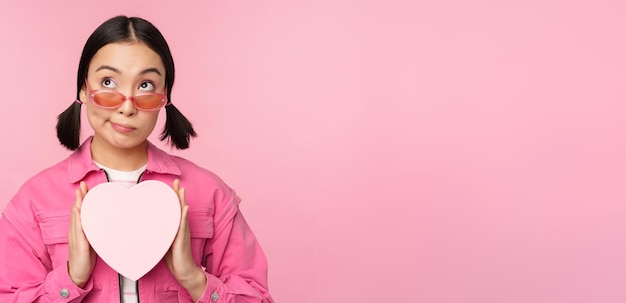 Linda garota asiática sorrindo feliz mostrando a caixa de presente do coração e olhando animada para a câmera em pé sobre fundo romântico rosa