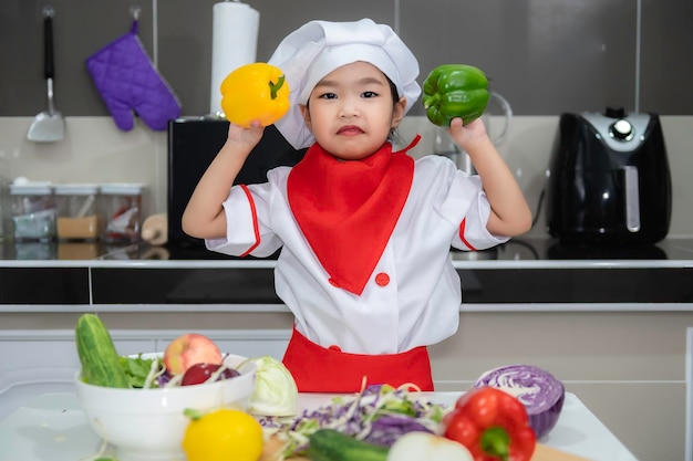 Linda garota asiática usa uniforme de chef com muita vegatable na mesa na sala da cozinhaFaça comida para o jantar Hora engraçada para crianças