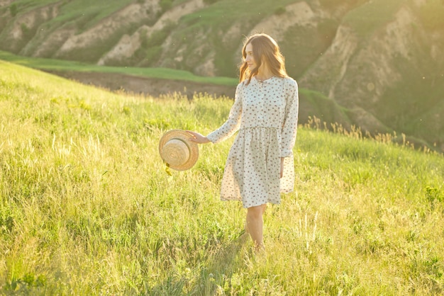 Linda garota atravessa as montanhas em suas mãos, segurando um chapéu de palha, apreciando os raios do pôr do sol.
