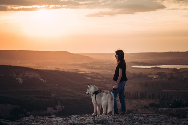 Linda garota brinca com um cão husky cinza e branco nas montanhas ao pôr do sol