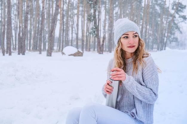 Linda garota caucasiana em um suéter branco e chapéu em um parque de floresta nevada bebendo chá quente ou chocolate de uma garrafa térmica. Garota vestindo roupas quentes de inverno durante as férias de Natal, Ano Novo