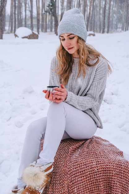 Linda garota caucasiana em um suéter branco e chapéu em um parque de floresta nevada bebendo chá quente ou chocolate de uma garrafa térmica. Garota vestindo roupas quentes de inverno durante as férias de Natal, Ano Novo