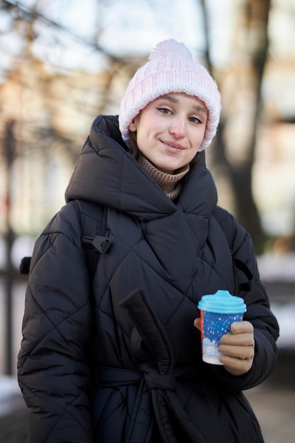 linda garota caucasiana em uma jaqueta preta e um chapéu de malha rosa, com uma xícara de café de papel.