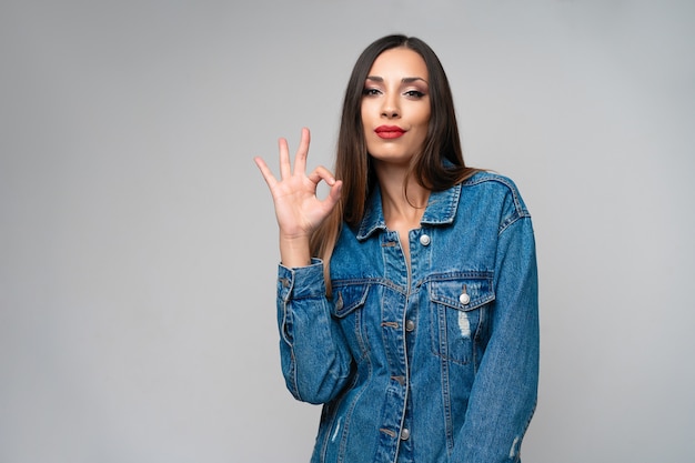 Linda garota caucasiana jaqueta jeans posando no estúdio, sobre fundo branco Morena bonito Denim moda lábios vermelhos compõem Jovem adulta em pé dentro de casa surpreendeu admirado Mostra ok gesto