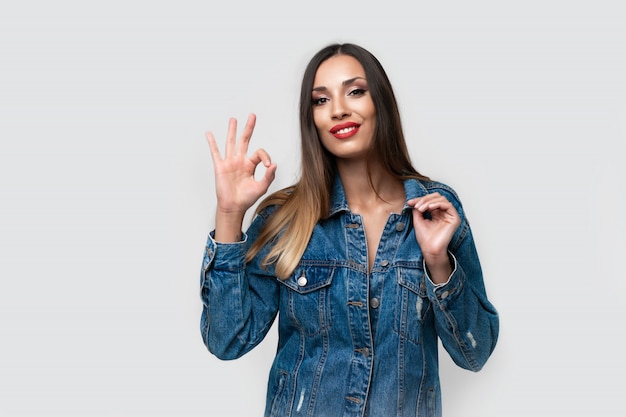 Linda garota caucasiana jaqueta jeans posando no estúdio, sobre fundo branco Morena bonito Denim moda lábios vermelhos compõem Jovem adulta em pé dentro de casa surpreendeu admirado Mostra ok gesto