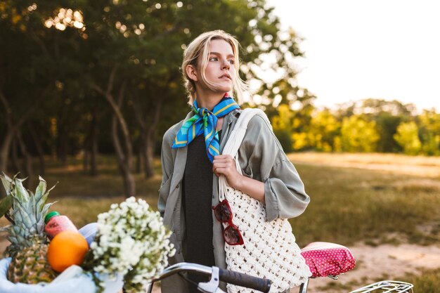 Linda garota com bicicleta e cesta cheia de flores silvestres e frutas olhando pensativamente de lado no parque