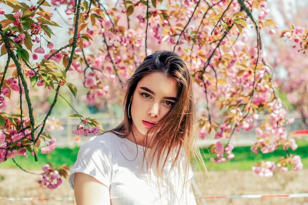 Linda garota com cabelo comprido aprecia a beleza da natureza na primavera, perto da florescente árvore de sakura.