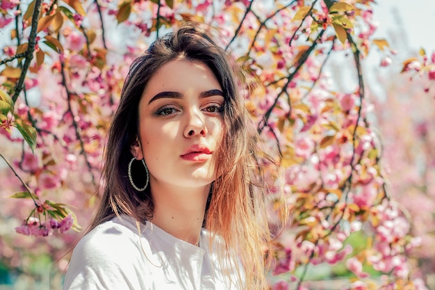 Linda garota com cabelo comprido aprecia a beleza da natureza na primavera, perto da florescente árvore de sakura.