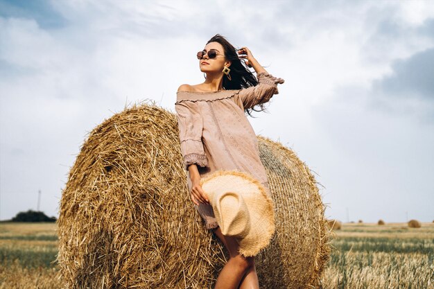 Linda garota com cabelo comprido em óculos de sol e chapéu de palha posando em um campo de trigo perto de fardos de feno. Morena feliz no vestido de verão