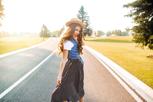 Linda garota com cabelo longo cacheado no chapéu está caminhando no sol na estrada, no pôr do sol. ela usa camiseta branca, gibão, saia longa, lábios vermelhos. ela parece ter gostado.