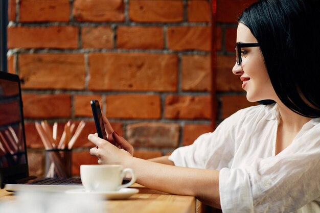 Linda garota com cabelo preto, usando óculos, sentado no café com laptop, celular e xícara de café, conceito freelance, retrato, cópia espaço, camisa branca.