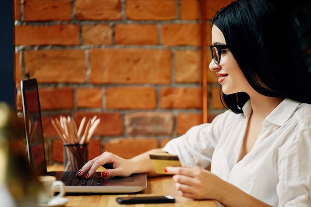 Linda garota com cabelo preto, usando óculos, sentado no café com laptop, telefone celular, cartão de crédito e xícara de café, conceito freelance, compras online, camisa branca.