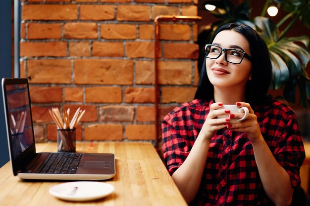 Linda garota com cabelo preto, usando óculos, sentado no café com o laptop e uma xícara de café, conceito freelance, retrato, vestindo a camisa vermelha.