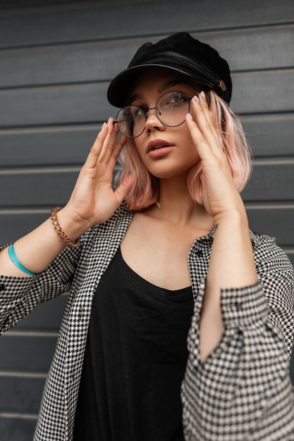 Linda garota com cabelo rosa e um boné em um streetwear estiloso com uma camisa e uma camiseta regata coloca os óculos e olha para a câmera