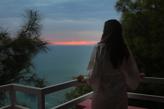 Linda garota com cabelos longos, de pé em um casaco branco na varanda do hotel com vista para o mar e o pôr do sol