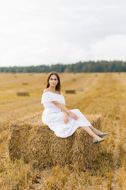 Linda garota com cabelos longos em um vestido longo branco, sentado em um palheiro em um campo