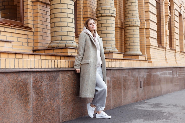 Linda garota com capuz biege elegante casaco cinza elegante e calças no fundo da parede de tijolos andando na rua