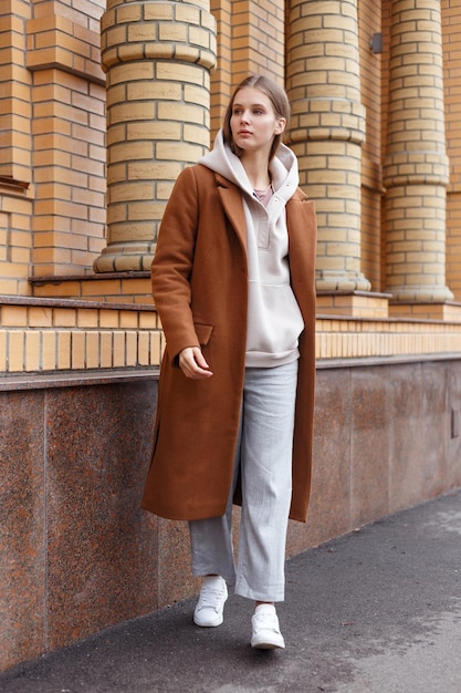 Linda garota com capuz elegante biege casaco de camelo marrom elegante e calças no fundo da parede de tijolos andando na rua