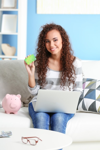 Foto linda garota com cartão de crédito, cofrinho e laptop no sofá