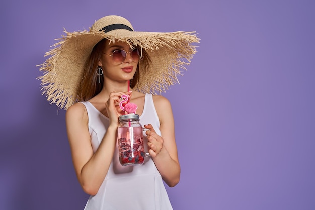 Linda garota com chapéu de palha e óculos escuros com um refrescante coquetel de verão no fundo roxo