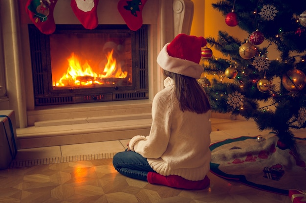 Linda garota com chapéu de Papai Noel sentada no chão e olhando para a lareira