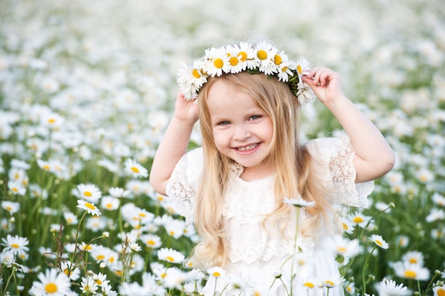 Linda garota com coroa de cabelos loiros e chamomilie no campo de camomila. retrato de criança menina bonitinha com buquê de flores de chamomiles em dia ensolarado de verão.