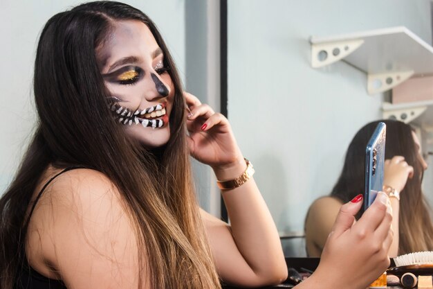 Linda garota com maquiagem de caveira de Halloween tirando selfies com seu telefone inteligente no quarto dela