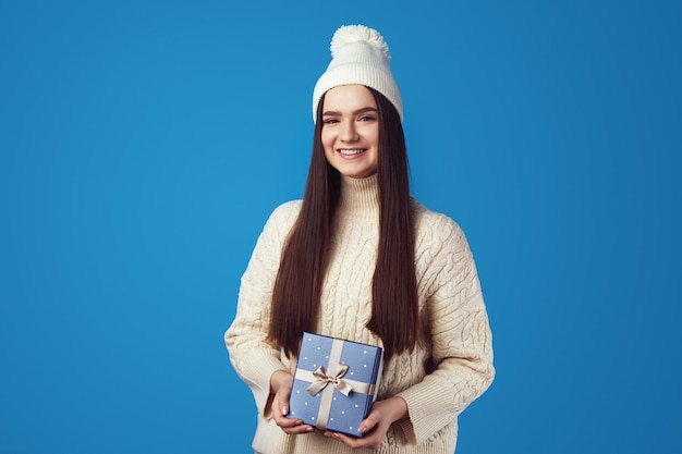 Linda garota com suéter branco e chapéu segurando uma caixa de presente azul embrulhada para