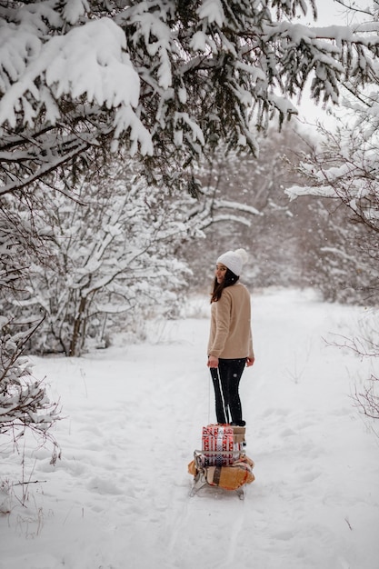 Linda garota com trenós, árvore de natal e presentes no inverno em um bosque nevado