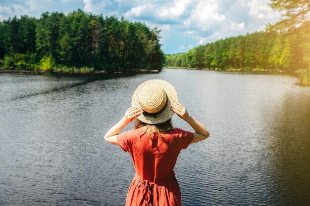 Linda garota com um chapéu de palha perto do lago. Natureza incrível de verão. Conceito de viagens. Inspiração para o desejo por viagens. Mulher de vestido de linho vermelho. Materiais ecológicos para roupas.