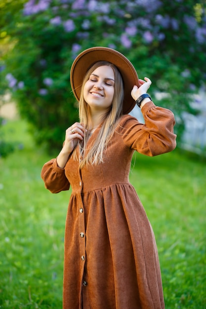 Linda garota com um chapéu marrom em pé perto de um arbusto de lilases. Mulher jovem perto de uma árvore florescendo.