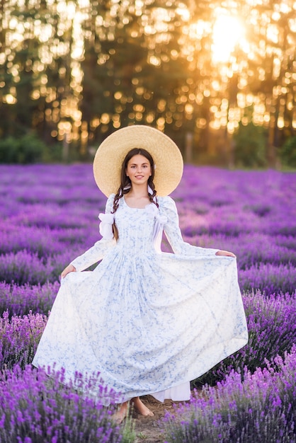 Linda garota com um grande chapéu e um vestido completo em um fundo de lavanda. Uma jovem modelo com tranças no cabelo. Foto de verão ao sol.