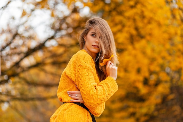 Linda garota com um moderno suéter de malha vintage amarelo em um parque de outono com folhagem de outono colorida