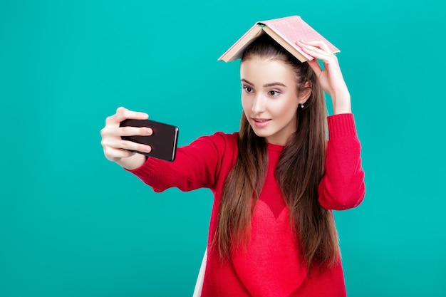 linda garota com um suéter vermelho lê um livro e usa um smartphone para selfie