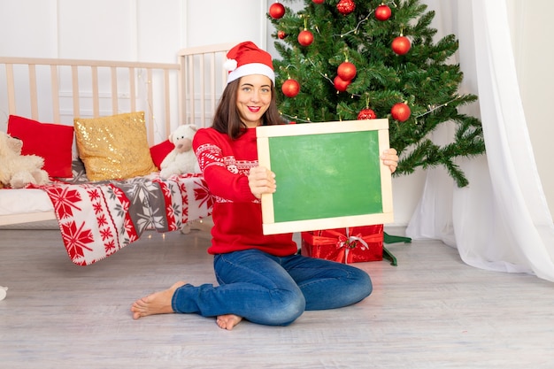 Linda garota com um suéter vermelho perto da árvore de Natal segura uma placa com espaço para texto