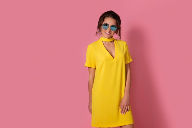 Linda garota com um vestido amarelo posando de óculos escuros, sorrindo no espaço rosa no estúdio