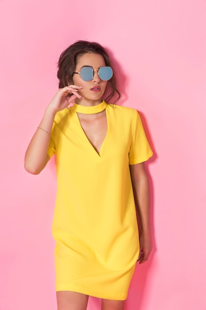 Linda garota com um vestido amarelo posando de óculos escuros, sorrindo no espaço rosa no estúdio