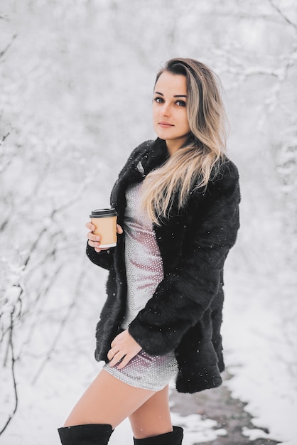 Linda garota com um vestido de casaco de pele e botas de cano altas no inverno na floresta com café nas mãos