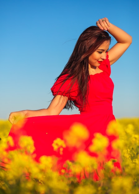Linda garota com um vestido vermelho, posando em um campo com canola. Mulher feliz na natureza. Temporada de primavera, dia quente.