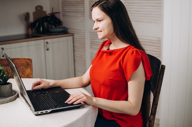 Linda garota com uma blusa vermelha trabalha em um laptop em casa O conceito de cursos on-line de trabalho remoto