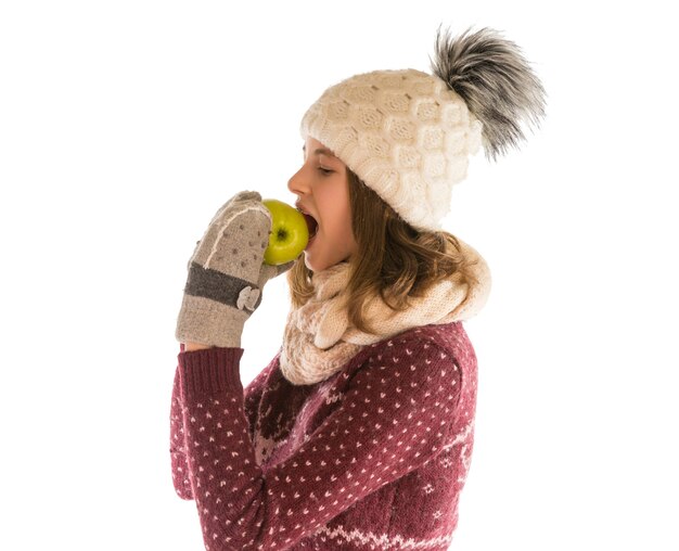 Linda garota com uma camisola quente, chapéu, lenço e luvas comendo uma maçã isolada no branco