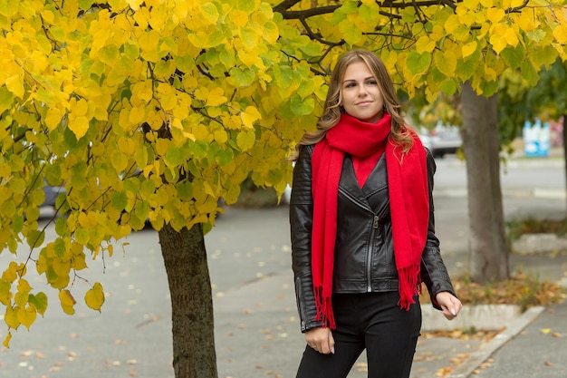 Linda garota com uma jaqueta de couro preta com um lenço vermelho perto de uma árvore com folhas amarelas