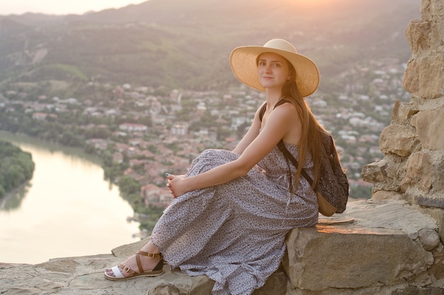 Linda garota com uma mochila e um chapéu largo, sentada na superfície do rio, das montanhas e da cidade abaixo