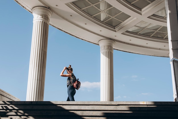 Foto linda garota com uma mochila em um ambiente urbano uma mulher caminha pela cidade em um dia ensolarado colunas clássicas da arquitetura da cidade