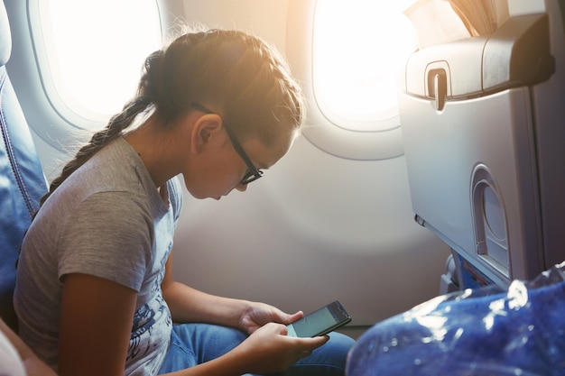 Foto linda garota com uma trança se senta em uma cadeira perto da janela da cabine da aeronave e se comunica nas redes sociais em um smartphone.