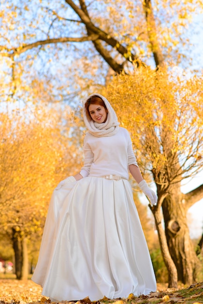 Linda garota com vestido de noiva longo branco, posando em um caminho rural entre árvores outonais na floresta