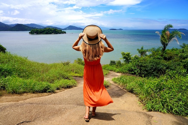 Linda garota com vestido de verão laranja segurando chapéu apreciando a vista da Costa Verde Brasil